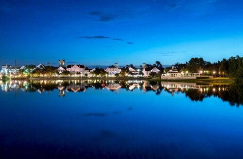 A serene lake at dusk, reflecting buildings and trees under a blue sky.