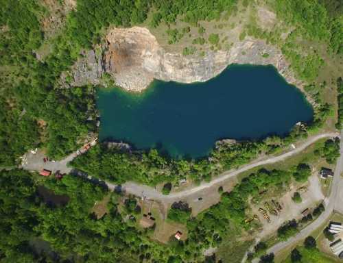 Aerial view of a blue lake surrounded by rocky cliffs and lush greenery, with a road and small structures nearby.