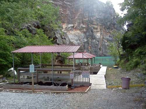 A wooden dock with gazebos beside a calm blue lake, surrounded by rocky cliffs and lush greenery.