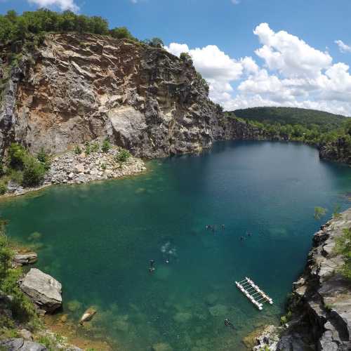 A scenic view of a rocky cliff surrounding a clear blue lake, with people swimming and a small dock visible.