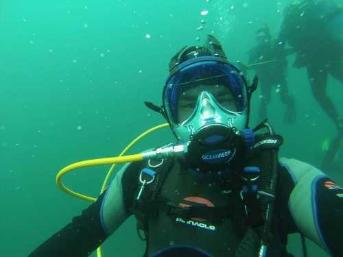 A diver in a wetsuit and mask underwater, with bubbles and other divers visible in the background.