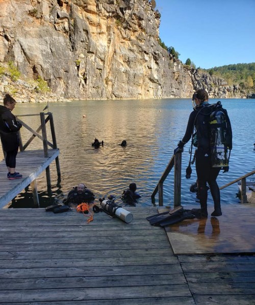 Divers preparing to enter the water near a rocky shoreline, with a wooden dock and spectators nearby.