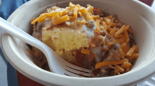 A bowl of cheesy cornbread topped with sausage, bacon, and melted cheddar, served with a plastic fork.