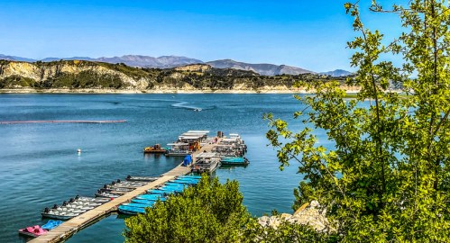 A scenic view of a calm lake with a wooden dock and colorful boats, surrounded by green trees and mountains in the background.