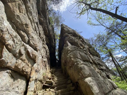 A narrow stone pathway leads through tall rock formations under a blue sky with scattered clouds and greenery.