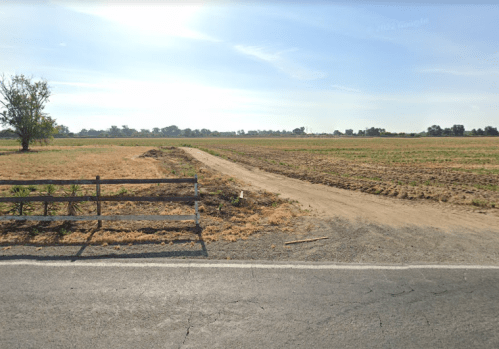 A dirt path runs alongside a grassy field, with a wooden fence and trees in the background under a clear sky.