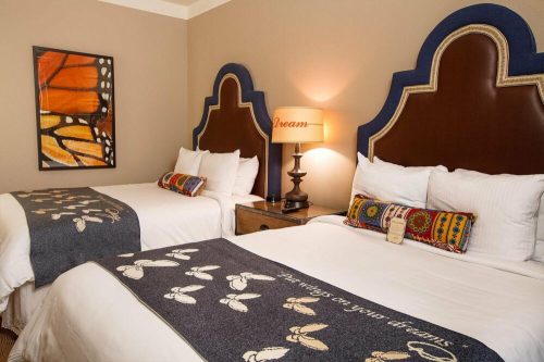 A cozy hotel room with two beds, decorative blankets, a lamp, and artwork on the wall.