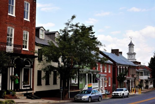 A charming street lined with historic buildings, trees, and a clock tower in the distance under a partly cloudy sky.