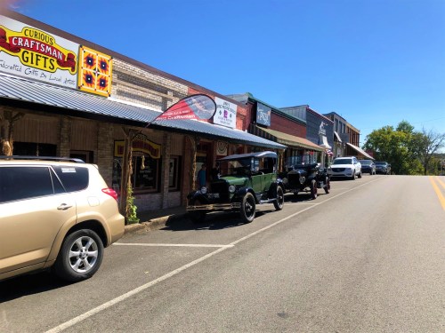A street lined with vintage cars and shops, featuring a sign for "Curious Craftsman Gifts" under a clear blue sky.