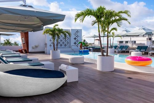 Rooftop pool area with lounge chairs, palm trees, and colorful inflatable floats under a blue sky.