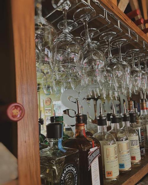 A wooden bar shelf displaying various liquor bottles and hanging wine glasses, with a sign that reads "Snaker."