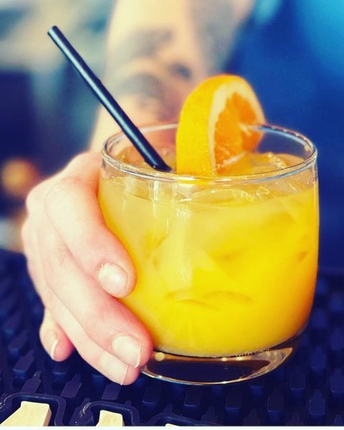 A hand holding a glass of orange cocktail with ice and a slice of orange, resting on a bar surface.