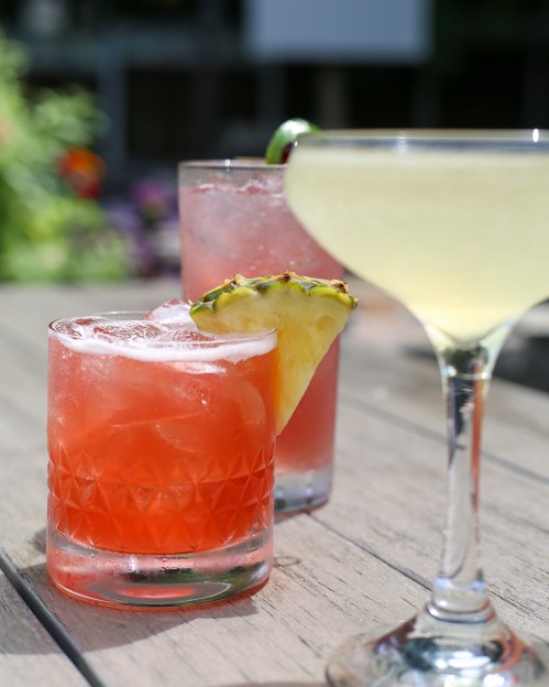 Three colorful cocktails on a wooden table: a red drink with ice and a pineapple slice, a pink drink, and a green cocktail.