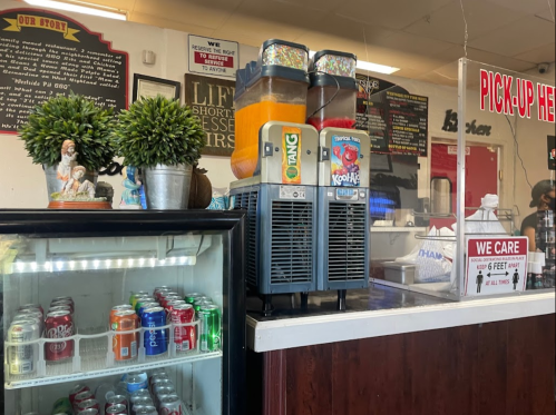 A colorful drink station with slushie machines, a soda fridge, and a sign reading "PICK-UP HERE" in a casual eatery.