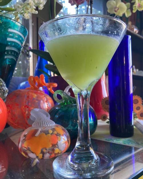 A green cocktail in a martini glass, surrounded by colorful decorative glass objects.