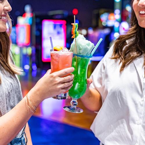 Two friends hold colorful cocktails with garnishes, smiling in a vibrant arcade setting.