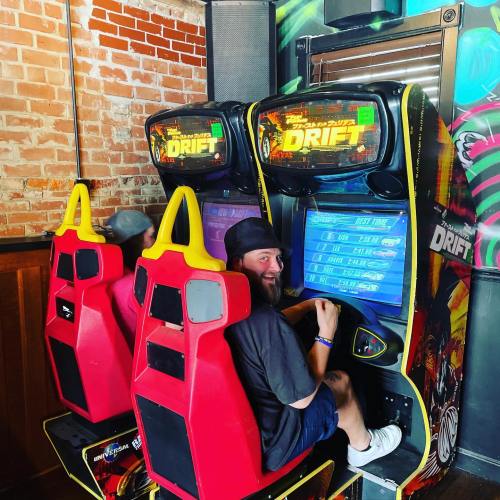 Two people playing a racing arcade game, seated in red and yellow chairs, with screens displaying "DRIFT" above them.
