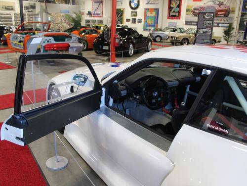 A white race car with an open door, showcasing its interior, in a car museum filled with various vehicles.