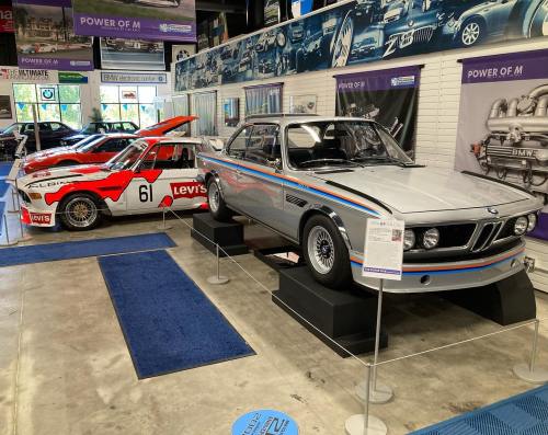 Two classic BMW race cars displayed in a museum, featuring vibrant colors and racing decals on a polished floor.
