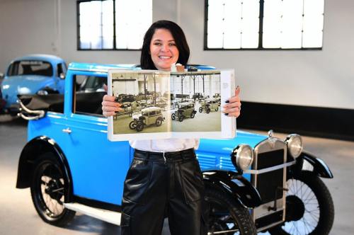 A woman holds an open book showcasing vintage cars, standing in front of a blue classic car in a museum.