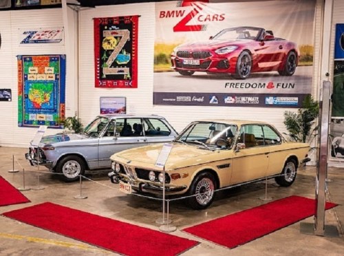 Two classic BMW cars displayed on a red carpet in a showroom, with a promotional banner in the background.