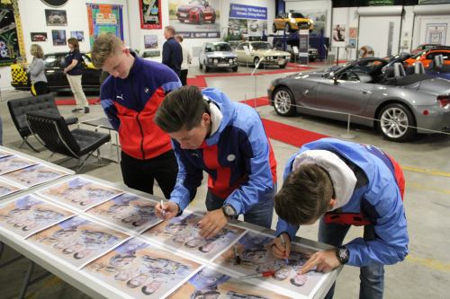 Three young men in jackets are signing posters at a car exhibition, surrounded by classic and modern cars.