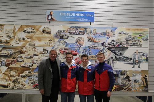 Three young men in colorful jackets pose in front of a mural depicting cars and racing scenes, with a man in a suit beside them.