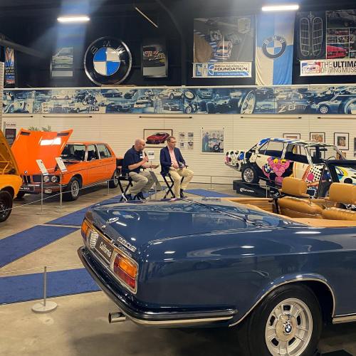 Two men sit in chairs discussing cars in a vintage car exhibit, featuring classic models and a BMW backdrop.