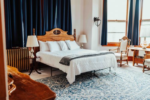 Cozy bedroom featuring a wooden bed, blue curtains, vintage chairs, and a patterned rug. Natural light from windows.
