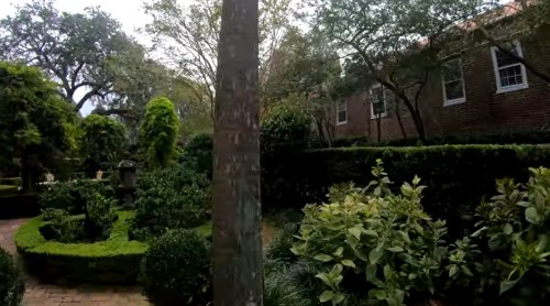 Lush garden with various plants, trees, and a brick building in the background under a cloudy sky.