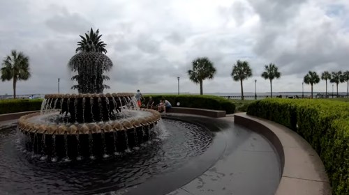 A decorative fountain with a pineapple design, surrounded by greenery and palm trees, under a cloudy sky.