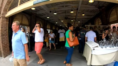 A bustling market scene with people browsing stalls, wearing summer clothing, and enjoying the atmosphere.