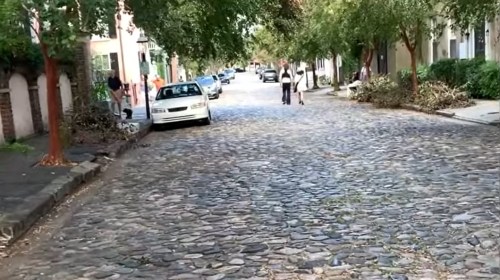A cobblestone street lined with trees, featuring parked cars and people walking in the distance.