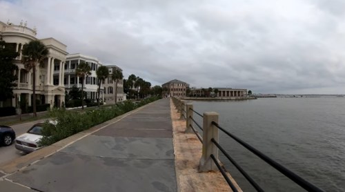 A waterfront view featuring a walkway, historic buildings, and palm trees under a cloudy sky.