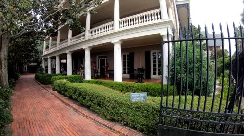 A historic building with a porch, surrounded by greenery and brick pathways, featuring a gated entrance.