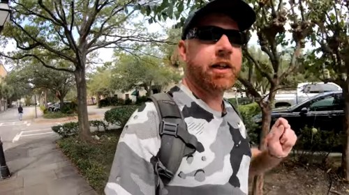 A man in sunglasses and a camo shirt walks along a tree-lined path, carrying a backpack.