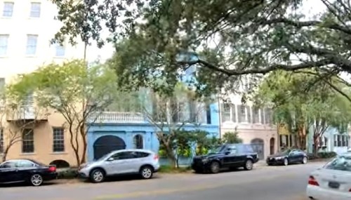 A street lined with colorful historic buildings and parked cars, shaded by trees.