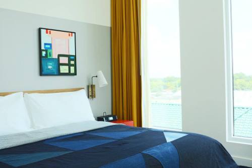 A modern bedroom featuring a bed with a blue quilt, a colorful wall art piece, and large windows with yellow curtains.