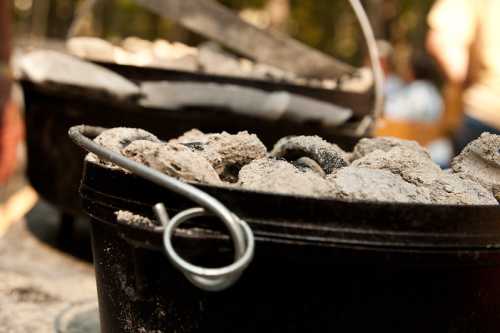 Close-up of a cast iron Dutch oven filled with coals, set outdoors with blurred background of people and trees.