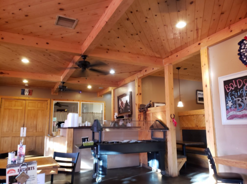 Cozy restaurant interior with wooden beams, tables, and a grill area, featuring warm lighting and rustic decor.