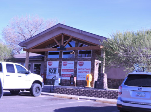Exterior of a sports bar with large windows, stone accents, and signage welcoming hunters. Vehicles parked in front.