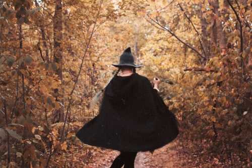 A person in a black cloak and witch hat walks through a forest of autumn leaves, creating a whimsical atmosphere.