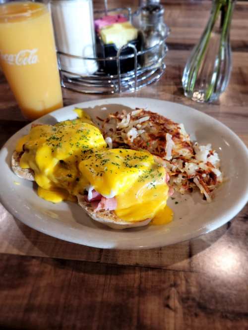 A plate of eggs Benedict topped with cheese, served with hash browns and a glass of orange juice in the background.
