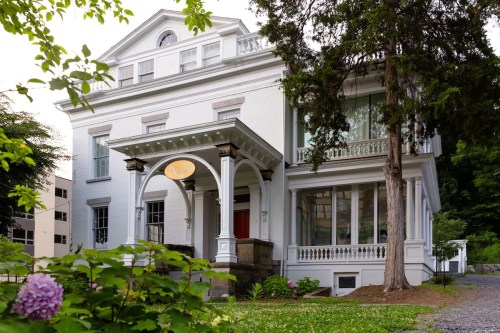 A large white historic house with a porch, surrounded by greenery and flowers, showcasing classic architectural details.