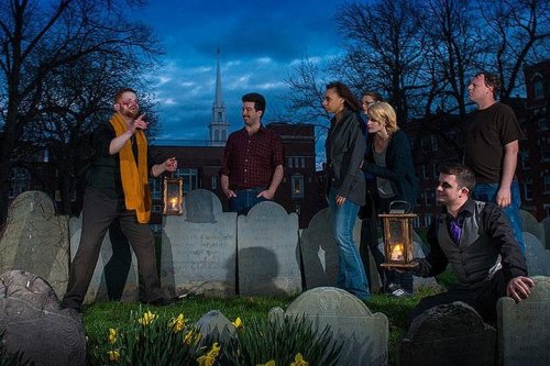 A group of seven people with lanterns gathered in a graveyard at dusk, listening to a storyteller.