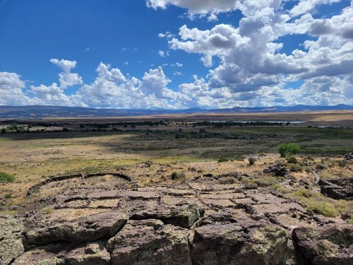 A vast landscape with rolling hills, a river, and a blue sky filled with fluffy clouds. Rocky foreground adds texture.