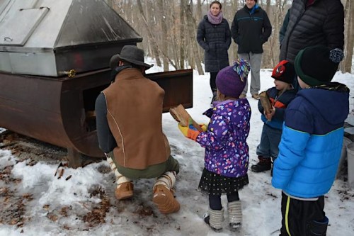 A person kneels by a metal stove in the snow, while children watch and hold firewood, surrounded by trees.