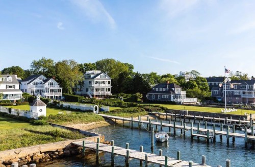 A scenic view of waterfront homes and a dock with a small boat, surrounded by lush greenery and blue skies.