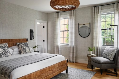 A modern bedroom featuring a woven bed, gray walls, large windows, and stylish decor with a cozy armchair.