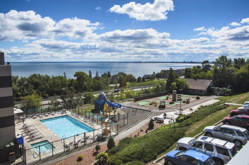 A scenic view of a pool area and playground by a lake, with cars parked nearby and a cloudy blue sky above.